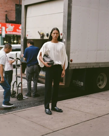 A model in a white blouse and black pants stands on a city sidewalk next to a moving truck with workers in the background.