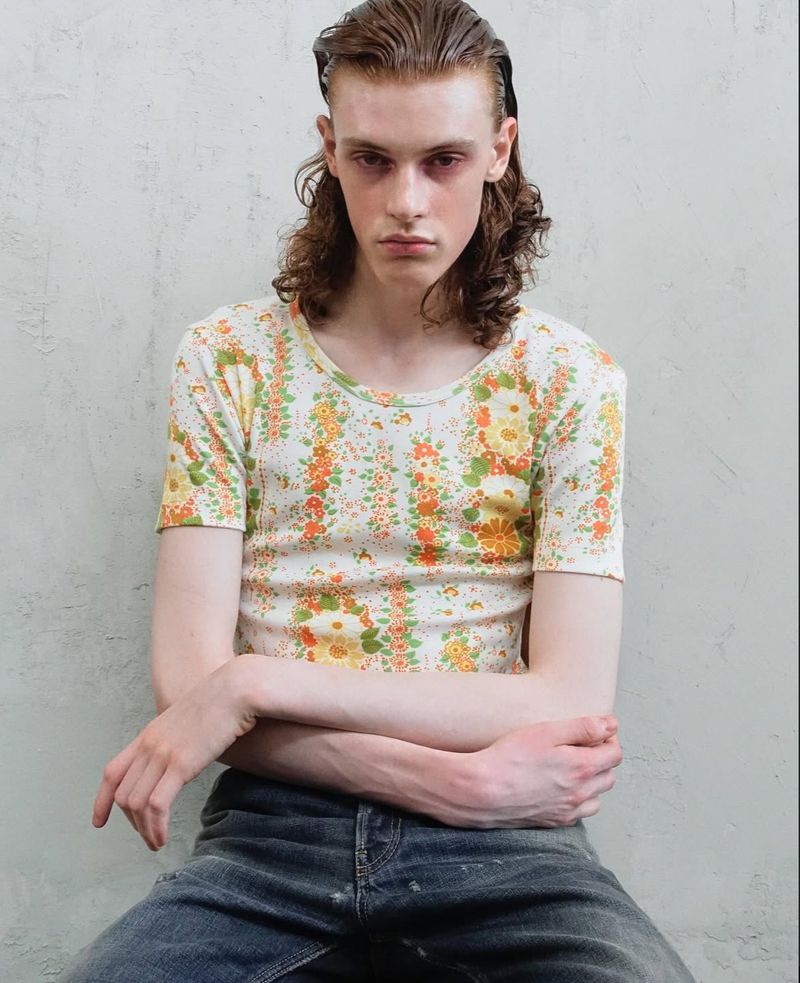 A young adult with shoulder-length wavy hair sits in a studio wearing a short-sleeved floral t-shirt and blue jeans, striking a nonchalant pose.