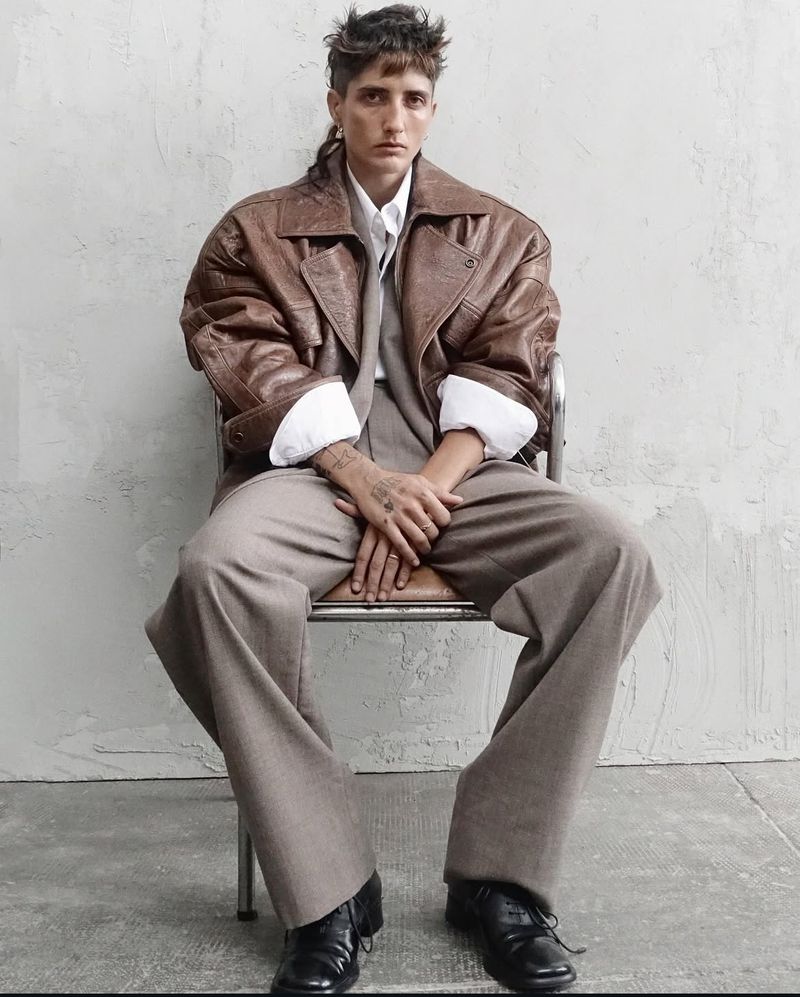 Adult woman in oversized brown leather jacket and wide-leg suit seated on a metal chair against a textured grey wall in a studio.