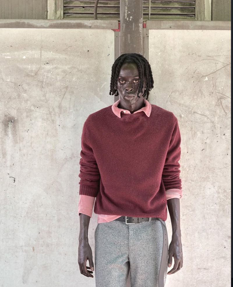 Portrait of a slender African male model in a maroon sweater and grey pants, posed against a textured white wall in an indoor setting.