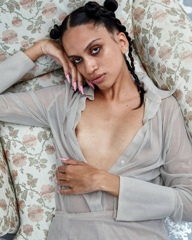 A woman with a pensive expression lying on a floral couch, wearing a semi-sheer shirt, her long pink nails visible.
