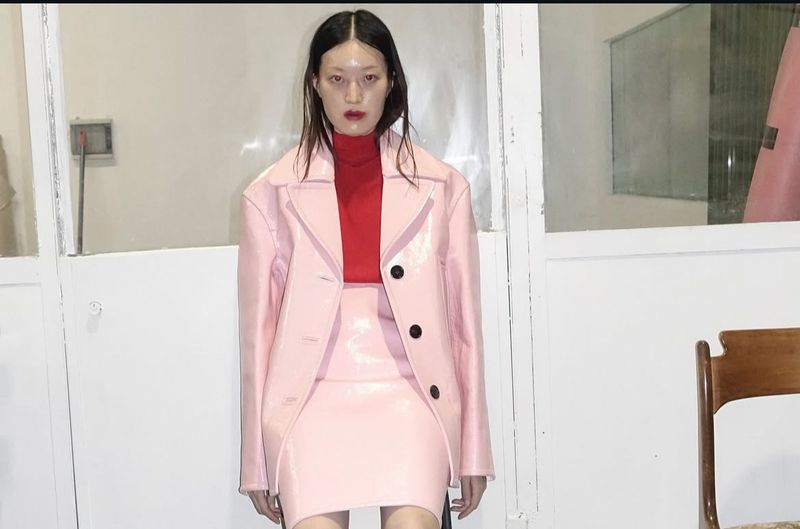A young adult woman poses in a photo studio, wearing a shiny pink skirt suit with a red turtleneck, against a stark white backdrop.