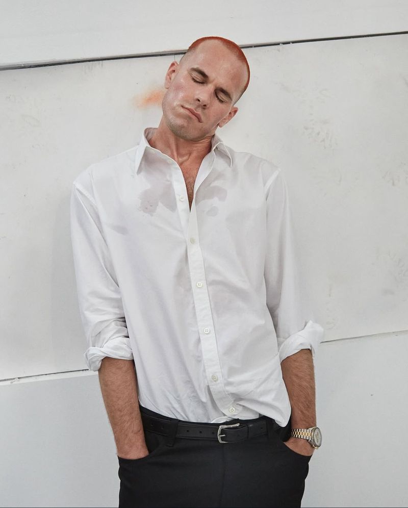 A man with red hair leans against a white wall, wearing a white button-down shirt and black pants, with his eyes closed in a studio setting.