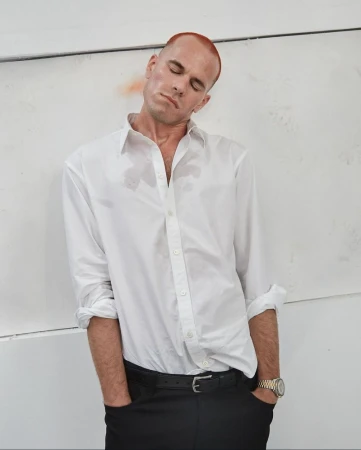 A man with red hair leans against a white wall, wearing a white button-down shirt and black pants, with his eyes closed in a studio setting.