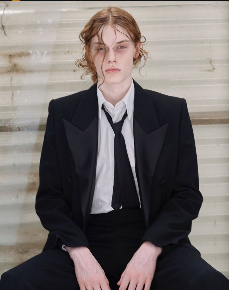 Portrait of a young adult male fashion model with long wavy auburn hair, wearing a black tuxedo with a white shirt and black tie. 