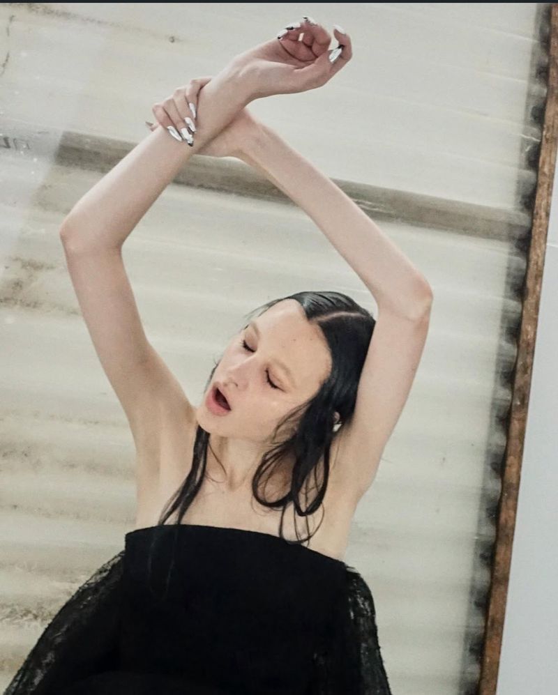 Portrait of a slender young woman with arms raised in a black top in front of a textured surface in a studio setting.