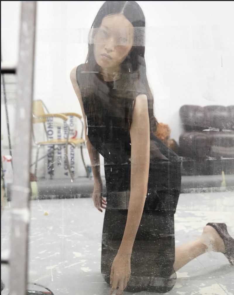 Model Martina Bergamini kneeling in a black sleeveless dress, photographed through a textured transparent pane in a studio setting.