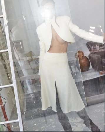 Model posing in a cream cropped jacket and matching midi skirt with front slit seen through reflective glass with camera flash flare.