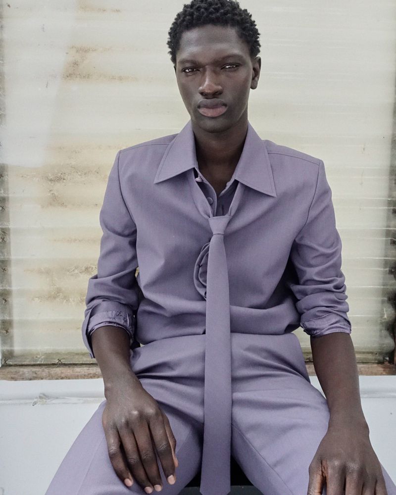 Portrait of a Black man in a light purple suit with a tie, sitting indoors with a neutral facial expression and textured wall background.