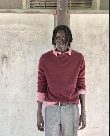 Model Modu Fall wearing a burgundy knit sweater, pink collared shirt, and grey trousers against a weathered industrial backdrop.