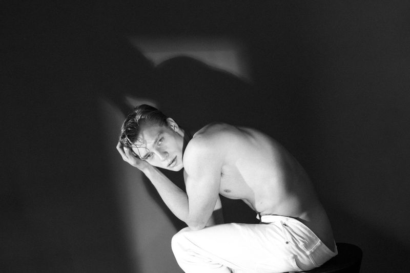 Monochrome portrait of a shirtless adult male sitting in front of a dark background with his hand resting on his head, looking at the viewer.