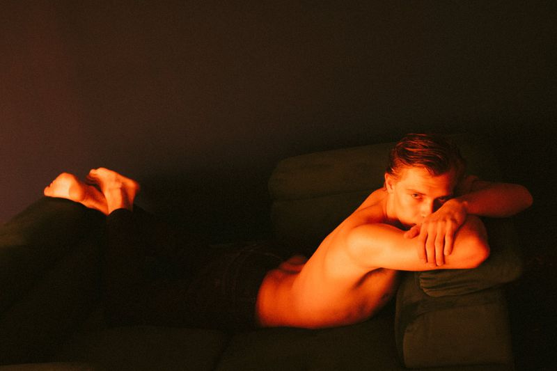 Bare-chested man lying on a couch illuminated with red lighting. He is resting his head on a pillow and has his hands folded.