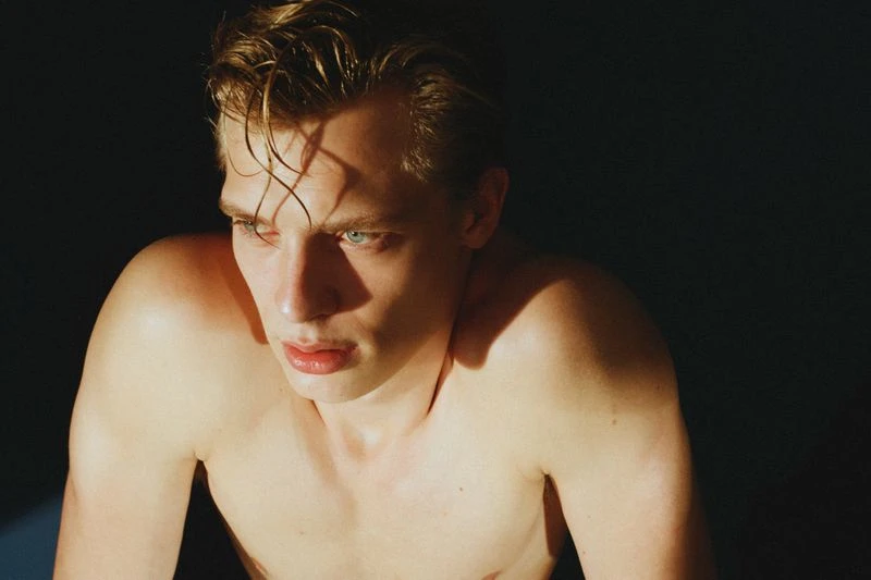 Close-up studio portrait of a blonde adult male with blue eyes, his torso is bare in dramatic spotlighting against a dark background.