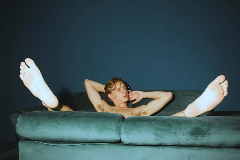 Shirtless blonde male adult with bare feet reclines on a teal velvet sofa against a dark blue wall in an indoor setting. 