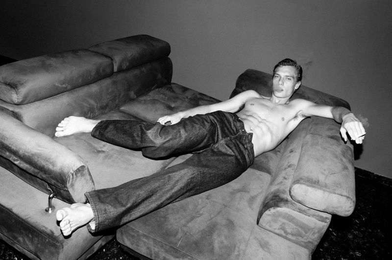 Black and white photo of a shirtless man in jeans lounging barefoot on a couch with an expressive face and hand gesture.