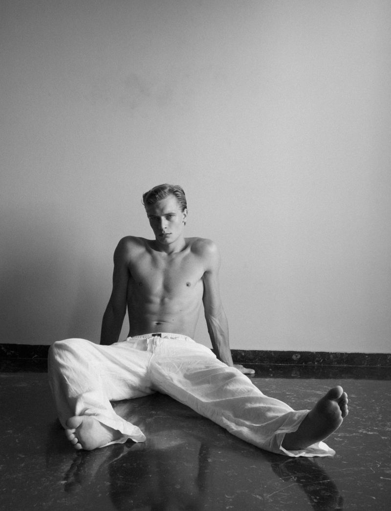 Black and white portrait of a shirtless adult male wearing white pants, sitting on a glossy floor against a plain wall.