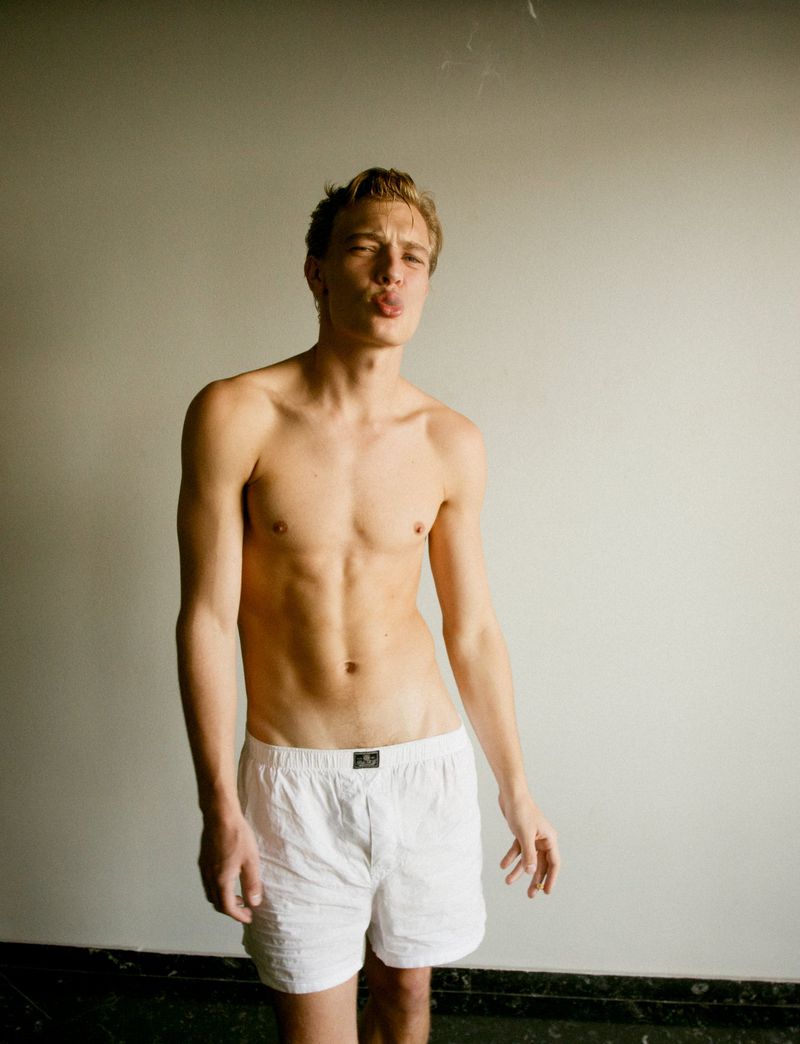 Male model poses shirtless in white boxer shorts against a plain background in a studio setting with soft, diffused lighting.