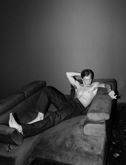 Monochrome portrait of a shirtless adult male reclining on a plush couch, holding a cigarette, set against a dark gray wall backdrop.
