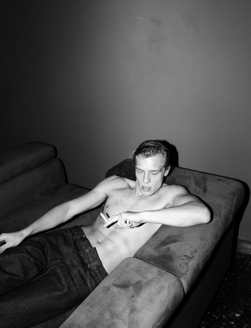 Shirtless young man in dark jeans reclining on a couch holding a cigarette in a black and white flash photograph.