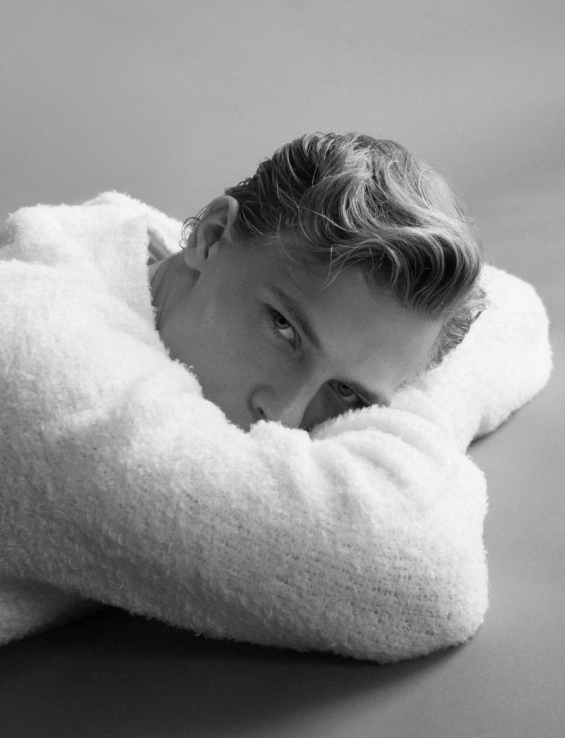 Black and white portrait of a young adult with fair skin and light hair, lying down, wearing a soft, textured, white sweater.