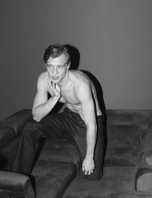A shirtless adult male seated on a dark velvet couch against a grey wall in a black and white studio photograph, posing with hand on face.