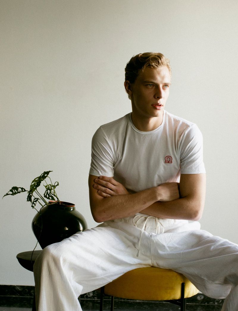 Blond man in white tee shirt and linen pants seated on a yellow stool with a plant nearby, posing in a bright, neutral indoor setting.