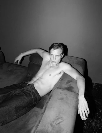 Shirtless young man reposing on a dark couch wearing denim jeans, holding a cigarette in a dimly lit interior setting.