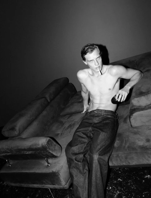 Monochrome portrait of a shirtless, slim adult male model in jeans smoking while seated on a retro couch against a dark background.