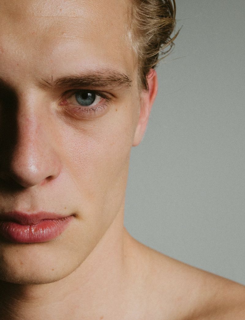 Striking close-up portrait of a young, fair-skinned adult male with light hair, set against a neutral gray backdrop in a photo studio.