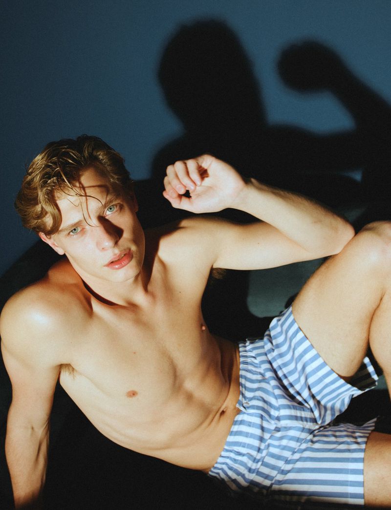 Blonde male model posing shirtless in striped shorts against a blue backdrop with shadow. Fashion and portrait photography.