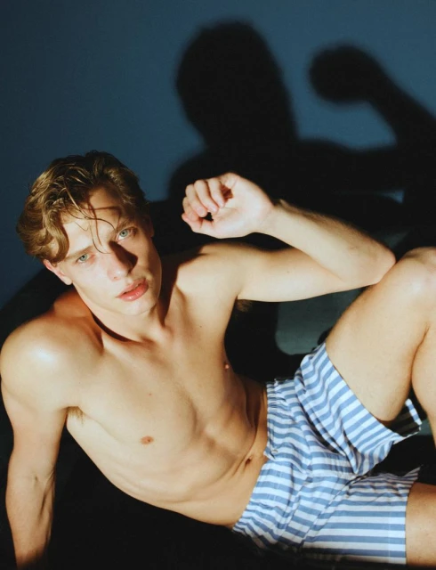 Blonde male model posing shirtless in striped shorts against a blue backdrop with shadow. Fashion and portrait photography.
