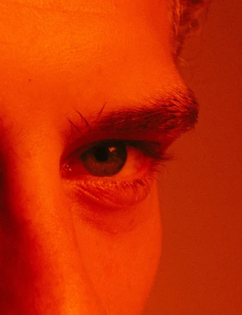 Close-up portrait of a young adult woman's eye and eyebrow in a dramatic orange-toned light, capturing detail and texture.