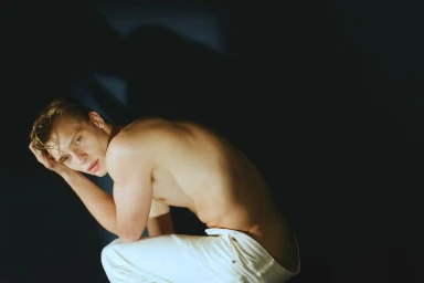 Shirtless man wearing white pants crouching against a dark background with a dramatic cast shadow.
