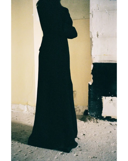 Person in a dark coat and dress posing indoors against a weathered wall, creating a somber and artistic fashion image.