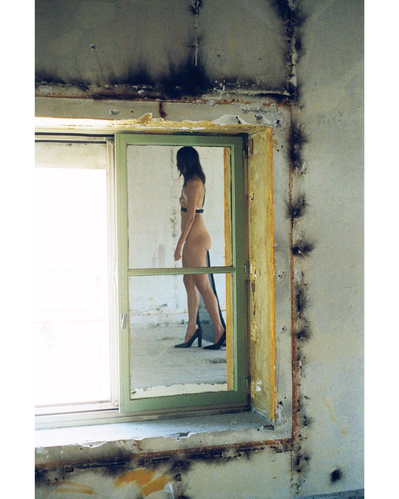 A woman in lingerie stands by a window in a worn building with peeling paint and black mold, with natural light from the window.