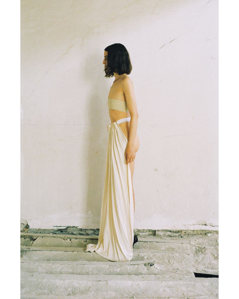Side profile of a person with short dark hair wearing beige strapless top and a beige draped garment, posing indoors against a white wall.