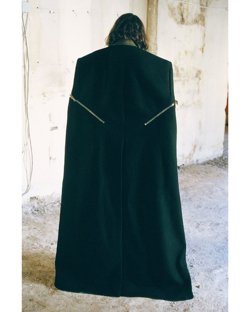 Person facing away from camera wears a black cape-like coat with exposed zippers against a weathered wall and floor in a minimalist setting.