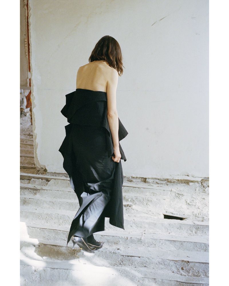 Woman in an abstract black strapless dress walks up grey stairs in an abandoned interior against a white wall in a fashion editorial.