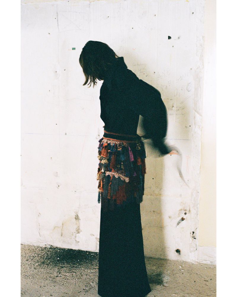 Model Akti Konstantinou standing in profile against a distressed white wall wearing a black long garment with a fringed textured hip wrap.