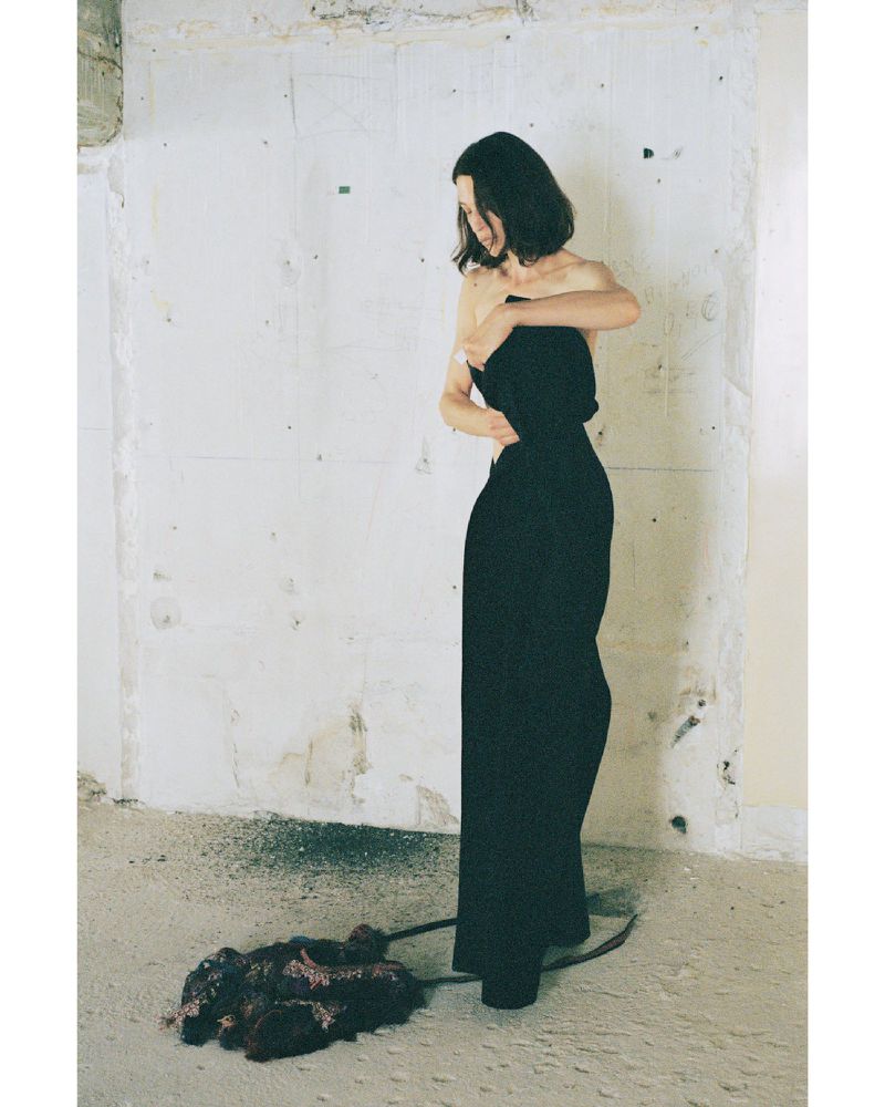 Model in black strapless dress standing against a distressed white wall with dark material on the floor.