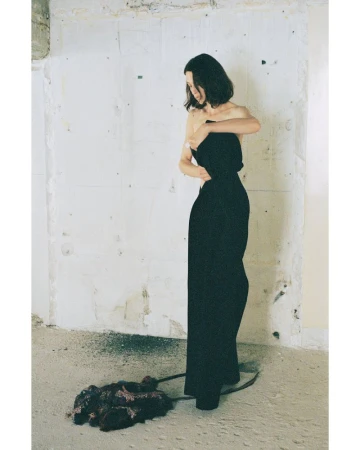 Model in black strapless dress standing against a distressed white wall with dark material on the floor.