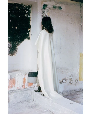Woman in flowing white dress stands in a worn, textured interior with visible wall damage, showcasing fashion and style in a raw environment.
