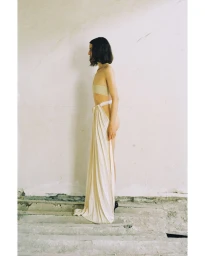 Model Akti Konstantinou standing in profile wearing a draped floor-length cream skirt and bandeau against a concrete wall.