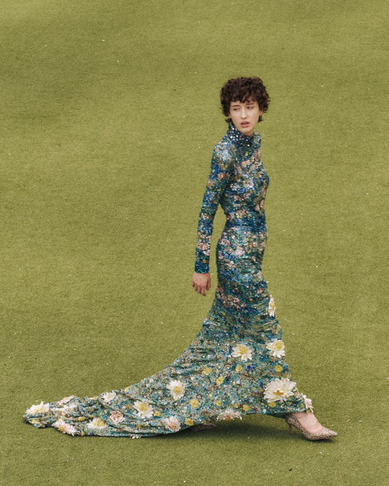 Woman in a floral dress walks across a green field. The dress has a long train and is covered in white and yellow flower details.