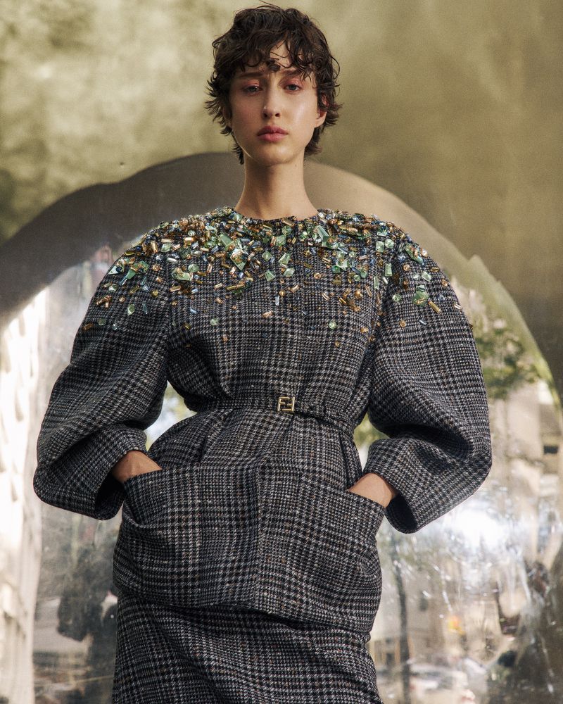A woman in a grey plaid embellished top and skirt is pictured in a medium shot against a blurred backdrop.