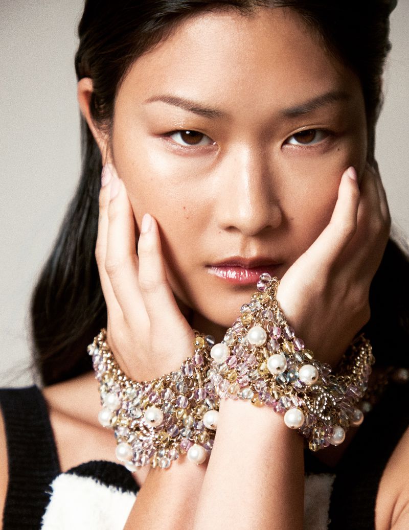Close-up of a young Asian woman with brown hair and fair skin, wearing pearl and gem encrusted bracelets holding her hands to her face.
