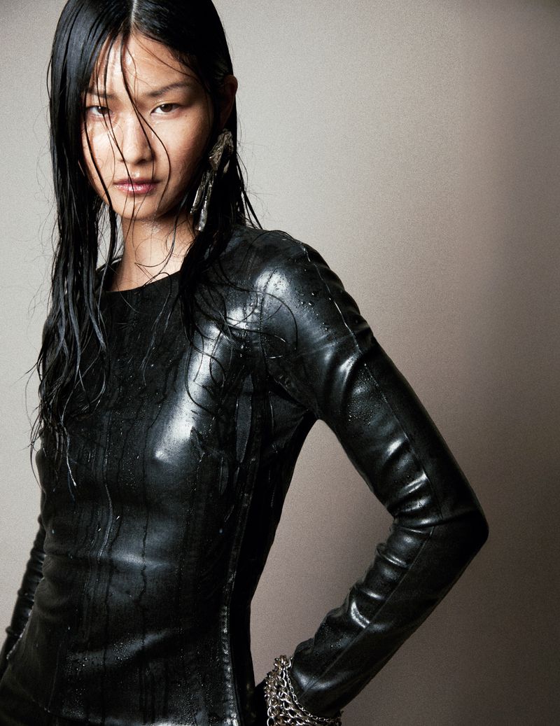 Portrait of Asian woman with wet, dark hair wearing black shiny dress and silver bracelet in a grey photo studio setting.