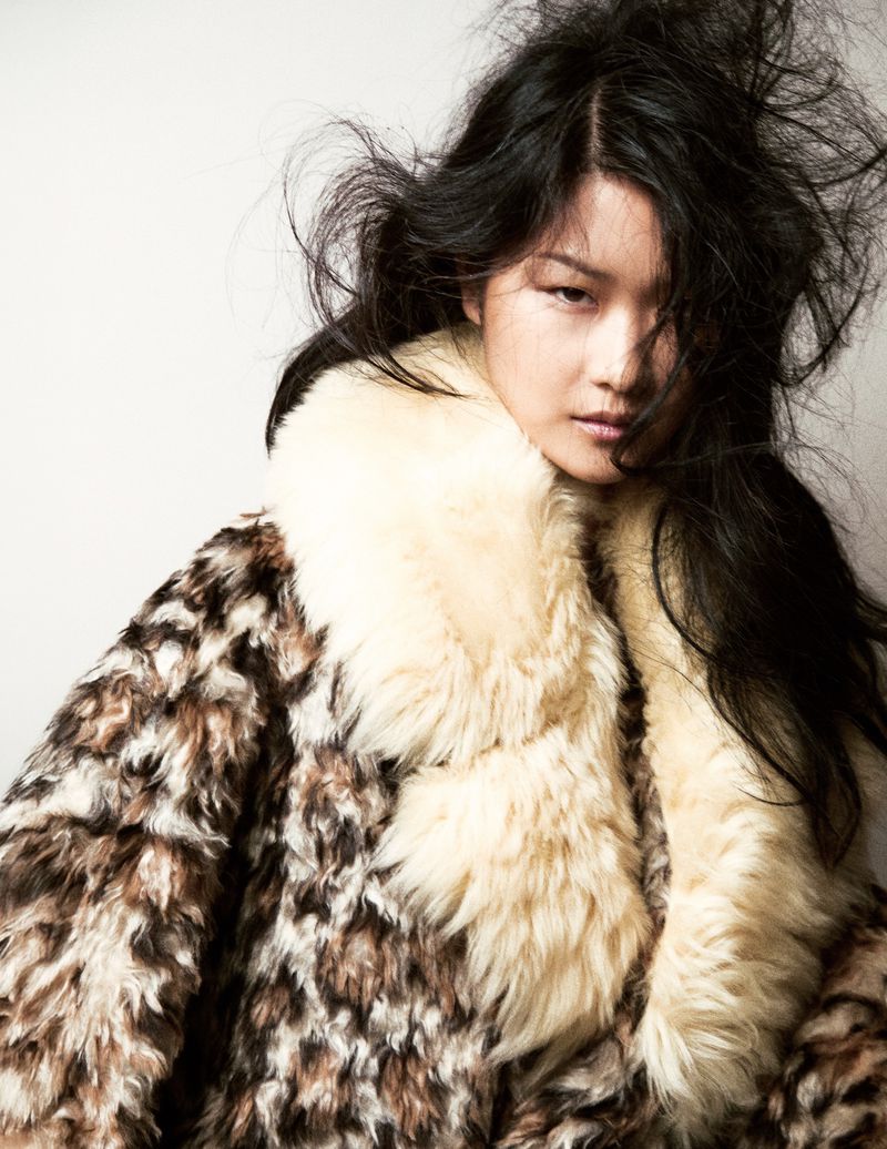 Portrait of a woman with dark hair wearing a patterned coat with fur collar against a white background in a fashion style.