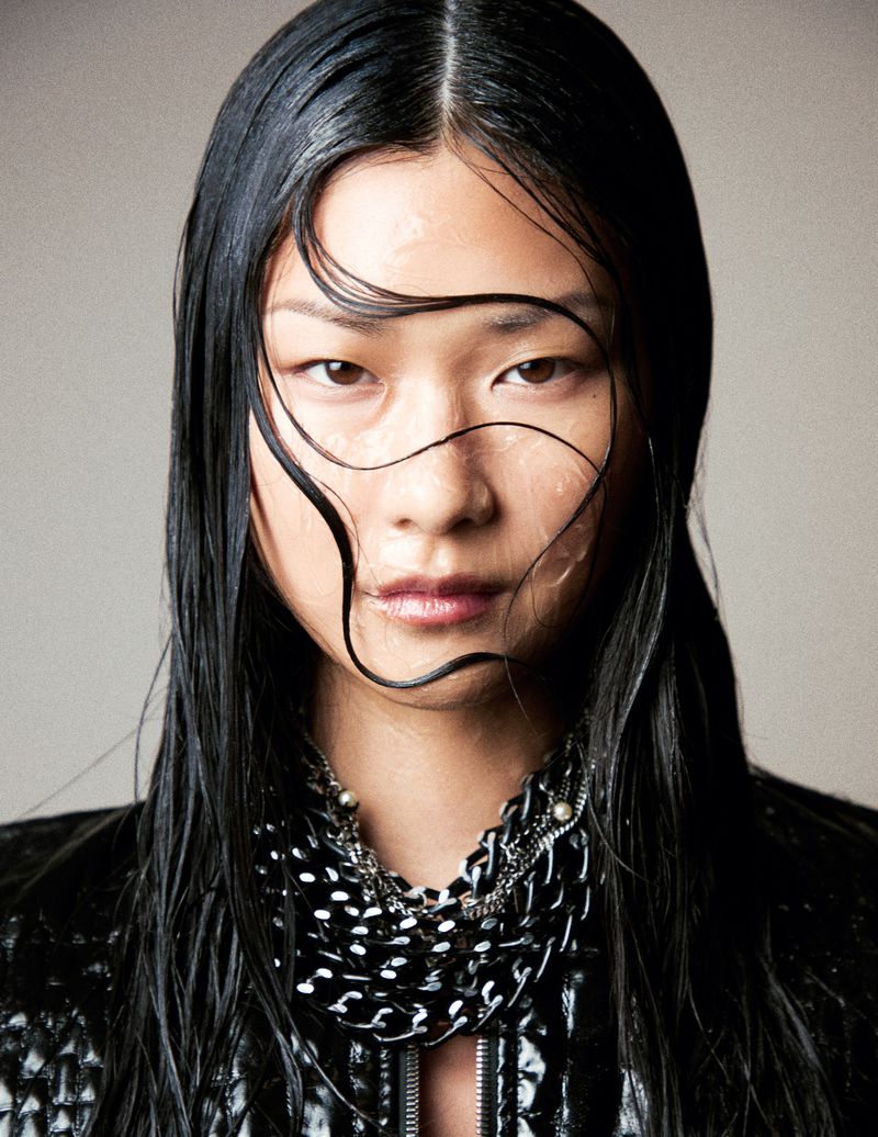 Portrait of a woman with wet, dark hair and a black jacket. She has light skin and is wearing a shiny black chain necklace.
