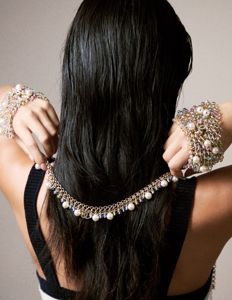 A model displays a gold necklace and pearl bracelets, with her black hair cascading down her back, set against a plain background.
